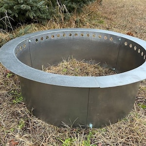 May include: A circular, silver-colored metal fire pit ring constructed from multiple curved panels joined by bolts. The top edge features a wide, flat rim with a series of evenly spaced circular ventilation holes along the inner circumference.