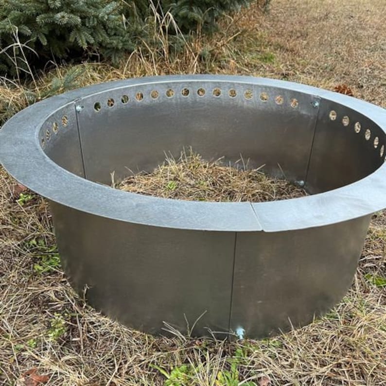 May include: A circular, silver-colored metal fire pit ring constructed from multiple curved panels joined by bolts. The top edge features a wide, flat rim with a series of evenly spaced circular ventilation holes along the inner circumference.