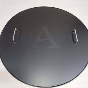 May include: Black round metal fire pit lid with two handles and the letter "A" engraved in the center.