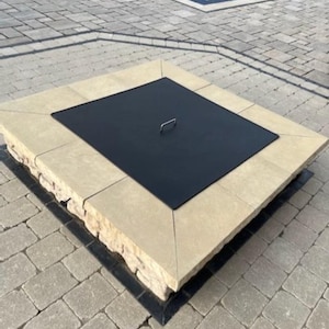 May include: A square fire pit with a beige stone surround and a black metal lid. The lid has a handle in the center.
