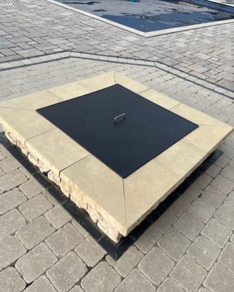 May include: A square fire pit with a beige stone surround and a black metal lid. The lid has a handle in the center.