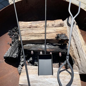 May include: A set of three black metal fireplace tools, including a poker, a shovel, and tongs. The tools are resting on a log of wood in front of a fire pit.