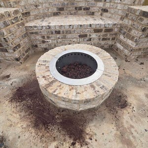 May include: A circular metal fire pit liner with a flat rim, installed within a round brick structure. The interior of the liner contains dark volcanic rocks. The fire pit is positioned in front of a curved brick bench wall made of light-colored bricks.