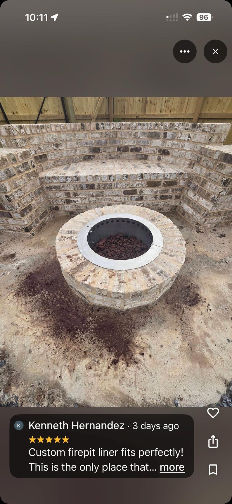 May include: A circular metal fire pit liner with a flat rim, installed within a round brick structure. The interior of the liner contains dark volcanic rocks. The fire pit is positioned in front of a curved brick bench wall made of light-colored bricks.