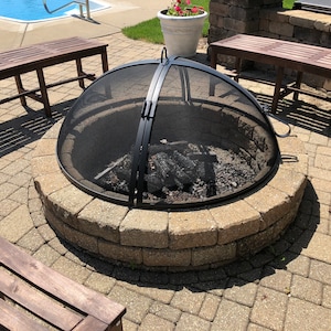 May include: A circular fire pit made of stacked stone, featuring a black mesh spark screen. The pit contains charred wood and sits on a paved patio. Two wooden benches are visible in the background, near a swimming pool.