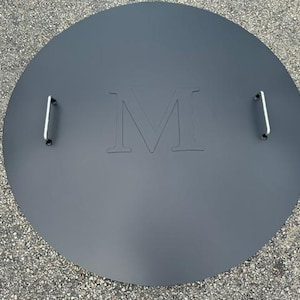 May include: A round, black metal fire pit cover with two handles and the letter "M" embossed in the center.