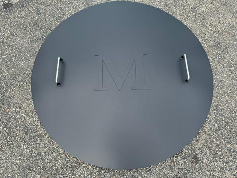 May include: A round, black metal fire pit cover with two handles and the letter "M" embossed in the center.