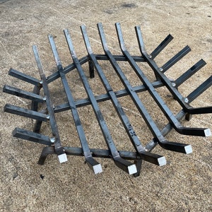 May include: A black metal fire grate with a circular shape and a grid pattern. The grate has a series of angled bars that form a ring around the center. The grate is designed to hold logs and other fuel for a fire pit.