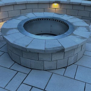 May include: A circular outdoor fire pit constructed from light gray concrete blocks with a metallic silver steel insert featuring a ring of ventilation holes along the top edge, set into a matching light gray concrete block patio and curved wall.