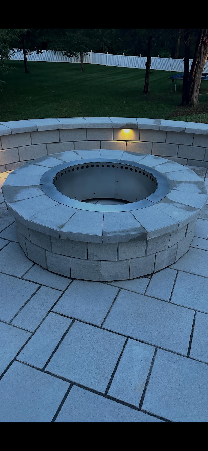 May include: A circular outdoor fire pit constructed from light gray concrete blocks with a metallic silver steel insert featuring a ring of ventilation holes along the top edge, set into a matching light gray concrete block patio and curved wall.