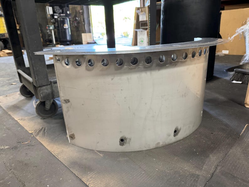 May include: A semi-circular, brushed metal component featuring a flat top flange and a row of evenly spaced circular perforations along the upper edge. The curved body includes several small, circular mounting holes positioned near the base.