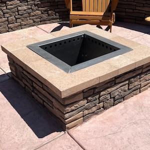 May include: A square fire pit made of stone with a black metal insert. The fire pit is surrounded by a stone wall and a wooden chair.