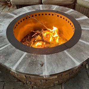 May include: A circular outdoor fire pit featuring a stone-tiled rim and a dark metal insert with a row of ventilation holes along the top edge. The center contains a burning fire with bright orange flames consuming small wooden branches.