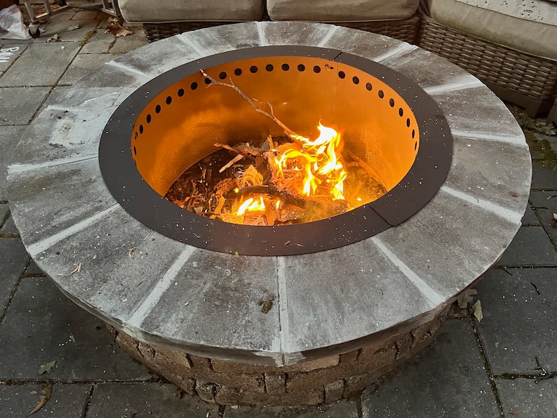 May include: A circular outdoor fire pit featuring a stone-tiled rim and a dark metal insert with a row of ventilation holes along the top edge. The center contains a burning fire with bright orange flames consuming small wooden branches.