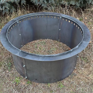 May include: A circular, dark gray metal fire pit ring constructed from multiple curved panels joined by visible bolts. The upper edge features a series of evenly spaced circular ventilation holes, and the structure has a wide, flat rim.
