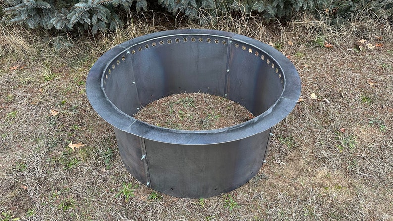 May include: A circular, dark gray metal fire pit ring constructed from multiple curved panels joined by visible bolts. The upper edge features a series of evenly spaced circular ventilation holes, and the structure has a wide, flat rim.