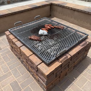 May include: A brick fire pit with a metal grill, featuring cooked sausages, steaks, and a foil-wrapped item. The pit is constructed of brown and tan bricks, set on a brick patio. The grill has handles.
