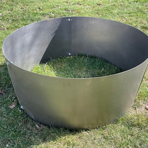 May include: A silver metal fire pit ring, sitting on a grassy lawn. The ring is round and has a slightly flared lip.