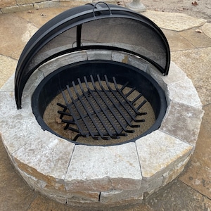 Op de afbeelding: Een zwarte metalen vuurkorf met een stenen omranding en een gaasdeksel. Het deksel is open en de vuurkorf is leeg.