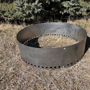 以下が含まれることがあります： 縁に穴が開いた丸い黒い金属製の焚き火台。リングは乾燥した草のベッドの上に置かれています。