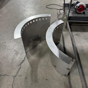 May include: Two curved, semi-circular metal panels made of brushed stainless steel. Each panel features a flat, horizontal top flange with a series of evenly spaced circular perforations along the edge and additional mounting holes on the vertical sides.