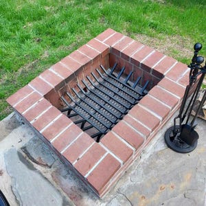 May include: A brick fire pit with a black metal grate and a set of black metal fireplace tools. The fire pit is surrounded by grass.