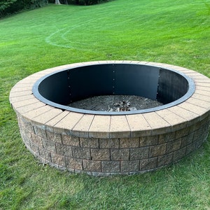 May include: A circular fire pit constructed from stacked tan and brown concrete blocks with a smooth, light-colored stone cap. The interior features a dark, curved metal liner secured with visible bolts, surrounding a base filled with dark gravel.