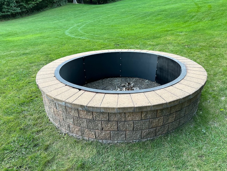 May include: A circular fire pit constructed from stacked tan and brown concrete blocks with a smooth, light-colored stone cap. The interior features a dark, curved metal liner secured with visible bolts, surrounding a base filled with dark gravel.