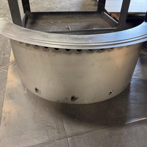May include: A semi-circular, silver-colored metal fire pit ring featuring a wide, flat top rim and a series of small, evenly spaced circular ventilation holes along the upper edge and near the base of the curved exterior wall.