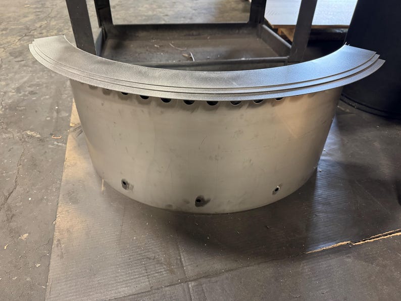 May include: A semi-circular, silver-colored metal fire pit ring featuring a wide, flat top rim and a series of small, evenly spaced circular ventilation holes along the upper edge and near the base of the curved exterior wall.