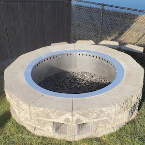 May include: A circular fire pit made of gray concrete blocks with a stainless steel ring around the top. The fire pit is filled with black gravel and has a metal grate at the bottom.