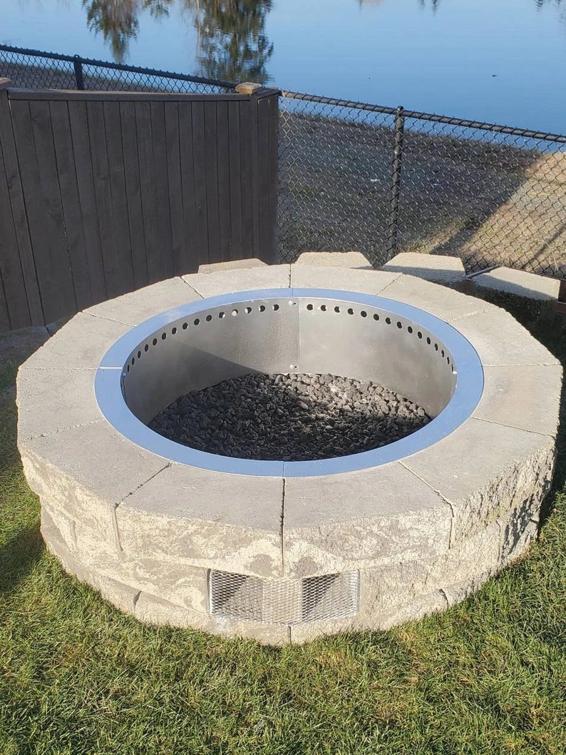 May include: A circular fire pit constructed from light gray concrete blocks with a smooth, metallic silver steel insert featuring a row of ventilation holes along the top edge. The interior is filled with dark gray lava rocks and a metal mesh vent.