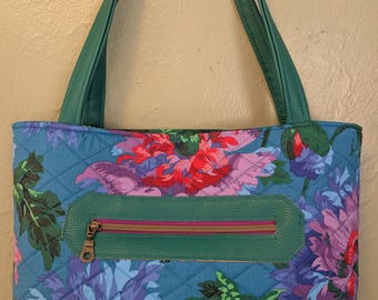 May include: A quilted tote bag with a floral design in blue, purple, and red. It has green leather handles and a green zippered pocket with a pink zip. The bag is designed for carrying items.
