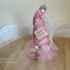 May include: A pink fabric party hat with white star patterns, a rhinestone heart, and a bow. The hat has a small patch with the number 31 and is adorned with a sheer, white veil and a large rhinestone button.