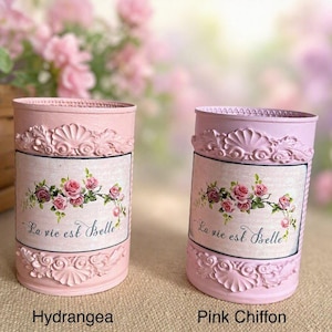 Könnte beinhalten: Zwei zylindrische Metallbehälter, einer in Hellrosa mit der Aufschrift Hydrangea und einer in Lavendel mit der Aufschrift Pink Chiffon. Beide haben ein zentrales rechteckiges Etikett mit Rosenillustration und dem Text La vie est Belle.