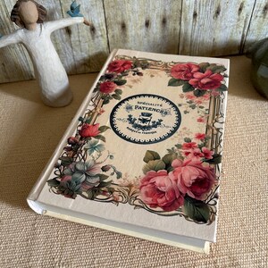 May include: A cream-coloured journal with a floral design on the cover. The design features pink roses, blue flowers, and green leaves. The journal has a gold-coloured border and a black circle in the centre with the text "SPECIALITE DE PATIENCES MARQUE DE FABRIQUE" in French.