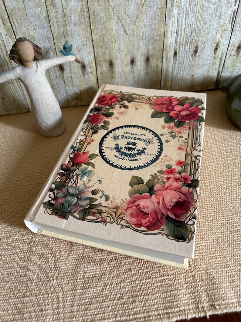 May include: A cream-coloured journal with a floral design on the cover. The design features pink roses, blue flowers, and green leaves. The journal has a gold-coloured border and a black circle in the centre with the text "SPECIALITE DE PATIENCES MARQUE DE FABRIQUE" in French.