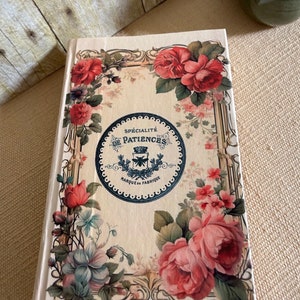 May include: A cream-coloured journal with a floral design featuring pink, red, and blue flowers. The cover has a black and white circular label with the text "Sp&eacute;cialit&eacute; de Patiences Marque de Fabrique".