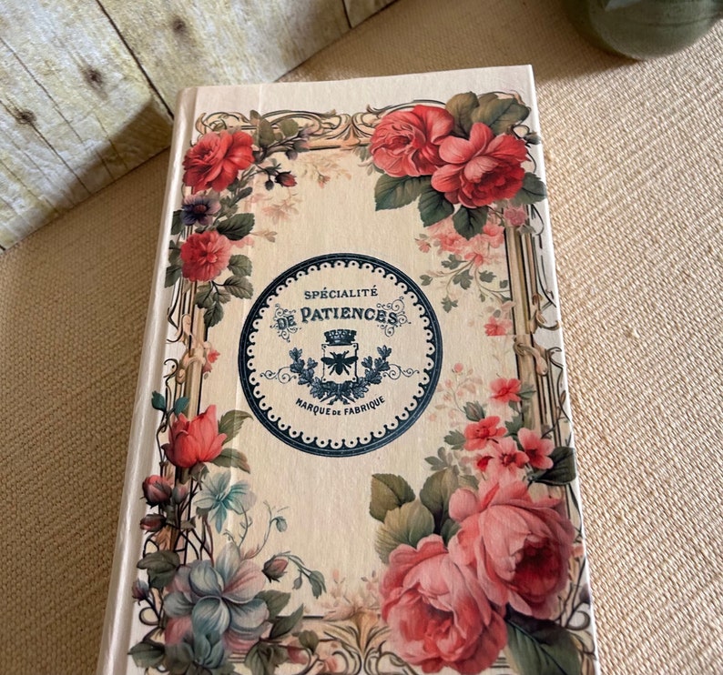 May include: A cream-coloured journal with a floral design featuring pink, red, and blue flowers. The cover has a black and white circular label with the text "Sp&eacute;cialit&eacute; de Patiences Marque de Fabrique".