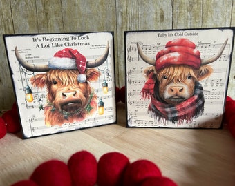May include: Two square wooden signs with Highland cow illustrations. One cow wears a Santa hat and a Christmas wreath, the other a red plaid scarf and hat. Both signs have sheet music backgrounds and holiday-themed text.