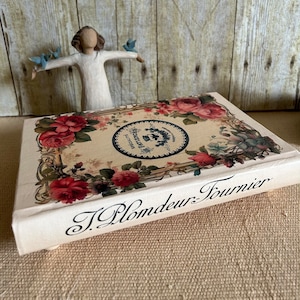 May include: A cream-coloured book with a floral design on the cover. The book is titled "J. Plomdeur-Tournier" and features a circular logo with the text "Specialite's - Parfumerie" in black and white.