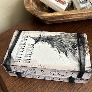 May include: A decorative box with the words "Rituals", "Curses & Spells", and "Potions" printed on it. The box features a black and white illustration of a witch and is trimmed with black lace.