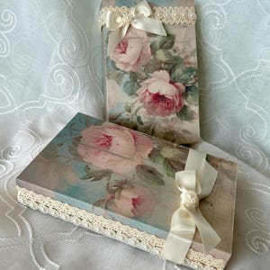 May include: Two decorative boxes with a floral design featuring pink roses and green leaves. The boxes are adorned with white lace trim and a satin ribbon tied in a bow.