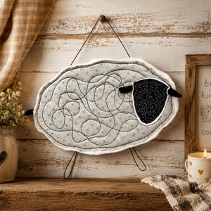 May include: A wall hanging shaped like a sheep, featuring a light gray body with a dark gray stitched swirling pattern and a black head with a white floral print. The sheep has a white fuzzy border and is suspended by a thin dark wire.