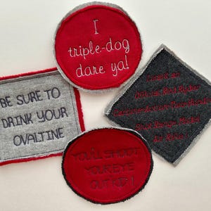 May include: Four fabric patches with embroidered text. One red circle reads I triple-dog dare ya!. One grey square reads BE SURE TO DRINK YOUR OVALTINE. One dark grey diamond reads I want an Official Red Ryder Carbine-Action Two-Hundred Shot Range Model Air Rifle!. One red oval reads YOU'LL SHOOT YOUR EYE OUT KID!.