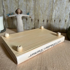 May include: A wooden book-shaped box with a white painted finish and the words "Remarkable Journey" printed on the side. The box has four wooden knobs on the bottom.