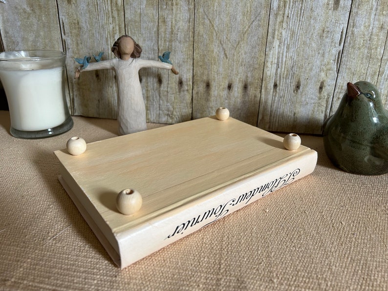 May include: A wooden book-shaped box with a white painted finish and the words "Remarkable Journey" printed on the side. The box has four wooden knobs on the bottom.