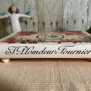 May include: A decorative wooden tray with a floral design and the text "J. Plomdeur-Fournier" printed on the front. The tray has four small wooden feet.