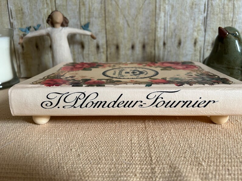 May include: A decorative wooden tray with a floral design and the text "J. Plomdeur-Fournier" printed on the front. The tray has four small wooden feet.