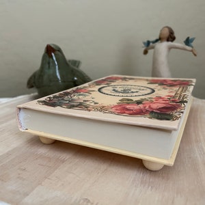 May include: A decorative box with a floral design and a white base. The box has four small feet and is made of wood. The box is closed and the top is decorated with a floral design.